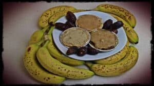 A number of ripe bananas are seen surround I white plate. Upon the plate, in aluminium tins are three delicious looking banana and date pies. The plate is also decorated with a few full dates.