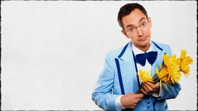 Myq Kaplan of “America’s got talent” is shown in a blue dress suit, holding a bunch of flowers.