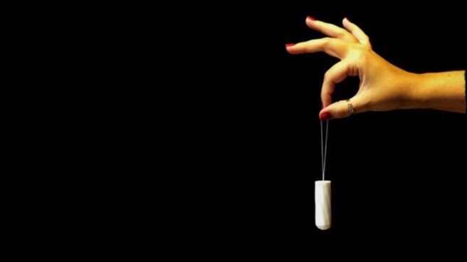 Against a dark background, from the right-hand side, a hand and forearm can be seen. The hand is holding the string of a dangling tampon between a finger and thumb.