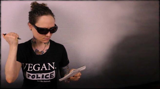 Emily Moran Barwick of Bite Size Vegan is shown in a black tee-shirt and sunglasses. On the front tee-shirt, in white, are the words: “Vegan Police”. She is about to make an entry into a pocket book.
