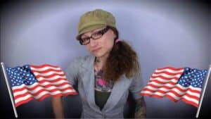 Emily Moran Barwick of Bite Size Vegan is standing between two billowing American flags. She has a wry expression on her face