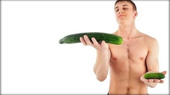 Naked man viewed from the waist up holding a small cucumber in one hand and a large cucumber in the other. The large cucumber is held higher than the small one