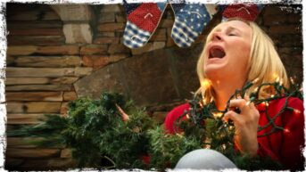 A person is shown with the branch of an artificial festive tree in one hand and festive lights in the other. Their head is back and they are letting out a scream of despair.