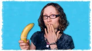 Emily Moran Barwick of Bite Size Vegan is shown holding a large banana. The banana has a condom rolled on to it. Emily is holding the fingers of her hand to her lips and showing surprize.
