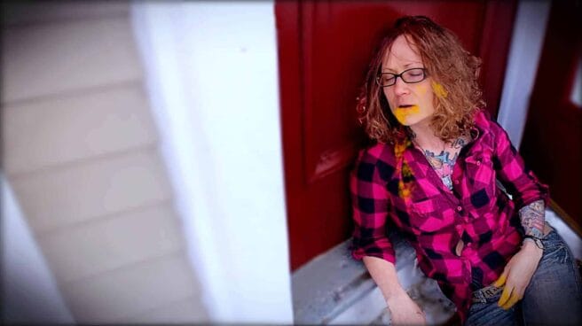 Emily Moran Barwick of Bite Size Vegan is shown in an apparent collapse, as if drunk. She is leaning up against a front door. There are marks around her mouth and chin as well as her fingers as she has been binging on cheese.