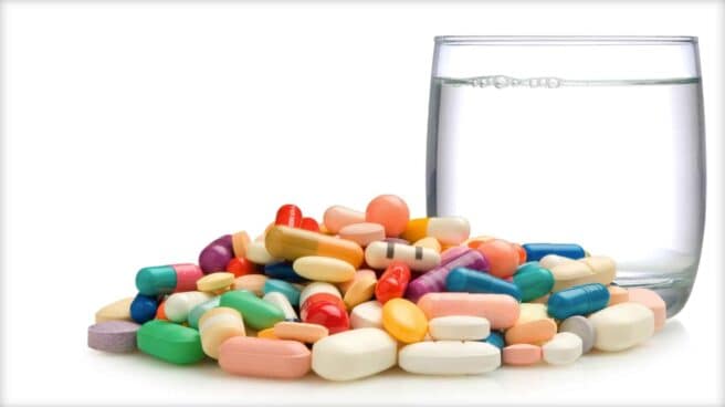 A large number of differing pills can be seen of different colors next to a glass of water.