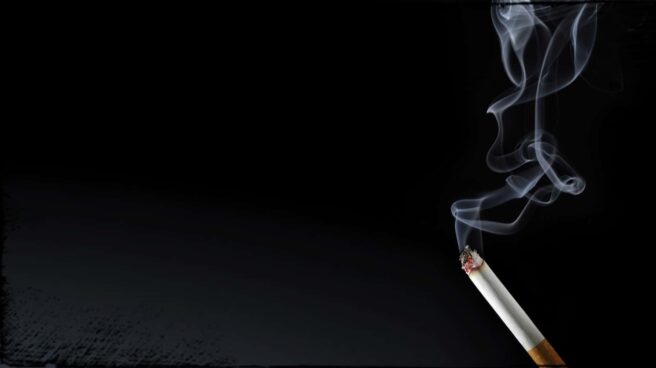 A lit cigarette with a trail of smoke rising into the air against a black background.