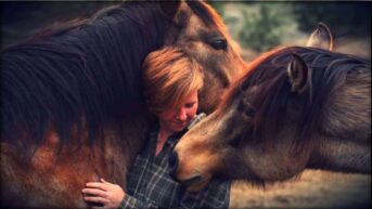 Author Ren Hurst embracing a horse who has nuzzled up to her and places his head over her shoulder, as a second horse brings her nose to Ren's.