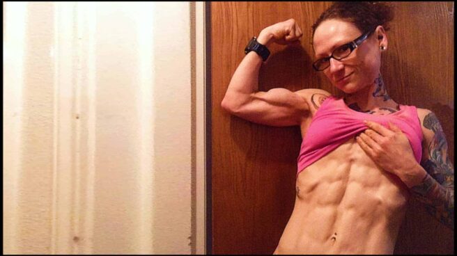 Emily Moran Barwick of Bite Size Vegan is shown from the waist up. Her right arm is curled, the muscles of her arm, clearly defined and taught. With her left arm, she is lifting up her shirt to reveal her wash-board abs and sculptured muscle definition.