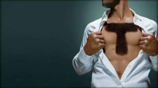 A man is shown from his chin to his waist. He is holding open the white shirt he is wearing, exposing his chest. Across the center of his chest, apparently rendered in chest hair, is a large capital letter T.