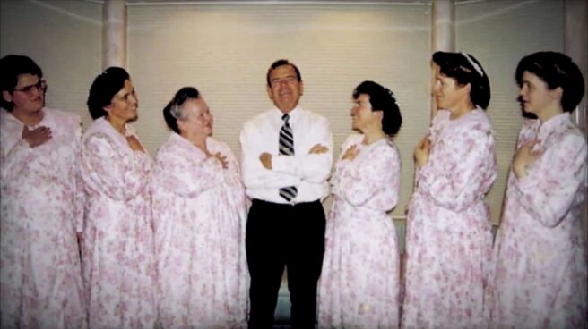 a man in black pants, white shirt and tie standing with his arms folded and smiling. On either side of him are 3 women dressed in similar dresses with pink flowers and similar hair do's gazing at him, 3 to his left and 3 to his right. The women to his right have their right hands over the hearts and the women to his left have their left hands over their hearts
