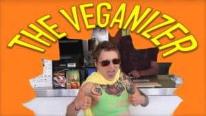 Emily Moran Barwick of Bite Size Vegan is shown dressed as the Veganizer superhero. The image makes it appear like she has punched her way through a wall. Across the top in large letters are the words “The Veganizer”