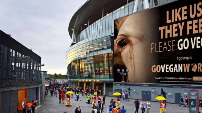 large outdoor city scene with large buildings with large billboard type ads showing a cow with tears running down it's face and the words Like us - they feel - go vegan. There are a lot of people walking around