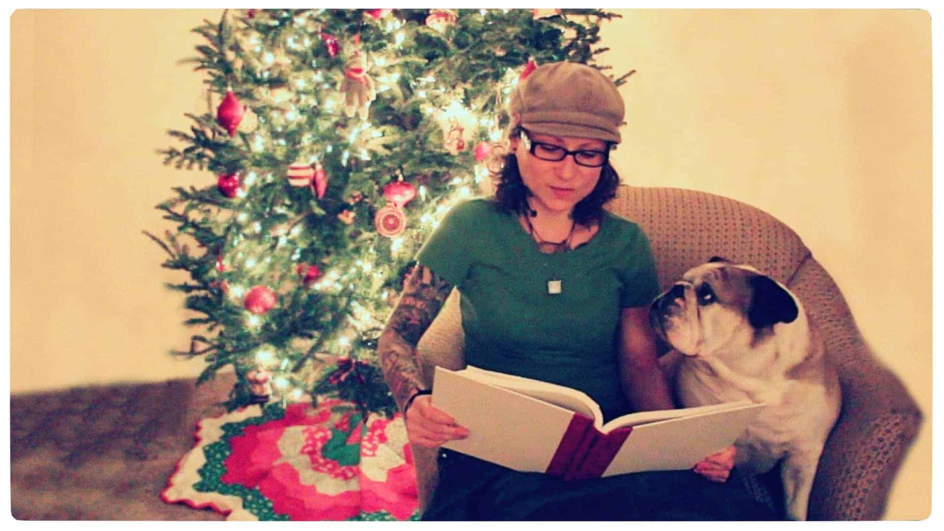 Emily Moran Barwick of Bite Size Vegan is shown sitting in a chair with her beloved bulldog, Ooby at her side and looking up at her. Emily is reading from a large book. Behind the tree is a fully decorated festive tree.