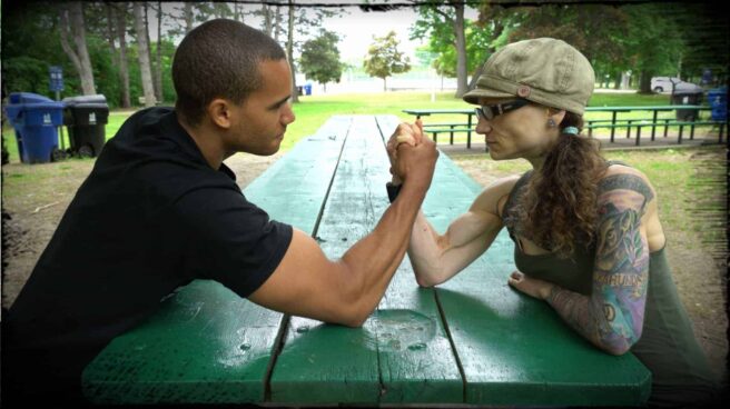Richard Burgess of Vegan Gains is shown facing off against Emily Moran Barwick of Bite Size Vegan in an epic arm wrestling showdown. They are facing each other on a park picnic bench, hands gripped, biceps taught, ready to begin.