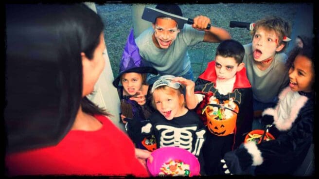 An adult is shown surrounded by children in Halloween costumes. The adult has a bowl of candy in their hand.