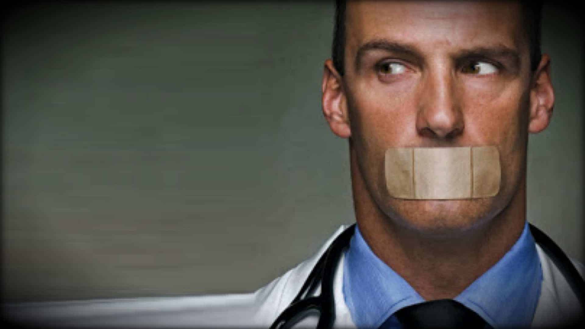 A person is shown in close up. They are wearing a white lab coat and a have a stethoscope around their necks. The are looking hard to one side whilst facing forward. A large self-adhesive plaster is fixed across their mouth, sealing it closed.