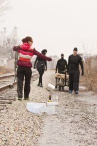 Emily is seen with a long chain looped over her shoulder at the side of the rail track as the individuals in black make their way towards her, pulling the trolley with them.  In the foreground the equipment that is to be used for the branding can be seen.