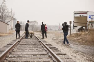 Three individuals clothed in black as well as a person holding a large video camera and Emily are shown in the distance standing around a rail road track.