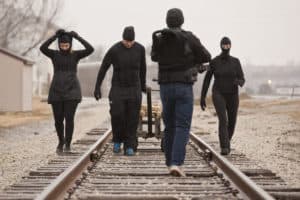 Three individuals clothed in black, two in masks are shown.  One is pulling the railroad truck.   A videographer is in front of them and is filming as the truck is pulled down the track.