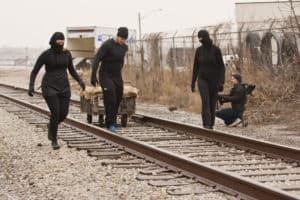 Three individuals clothed in black, two in masks are shown.  One is pulling a small four wheel trolley.   The trolley is approximately 5 feet by 3 feet and has a wooden top that is covered in rough canvas.  A videographer is at the side of the track and is filming as the truck is pulled down the centre of the railroad track. 