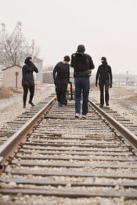 Three individuals clothed in black, two in masks are shown in the distance. One is pulling the railroad truck. The videographer has their back to the camera and is filming the truck scene.