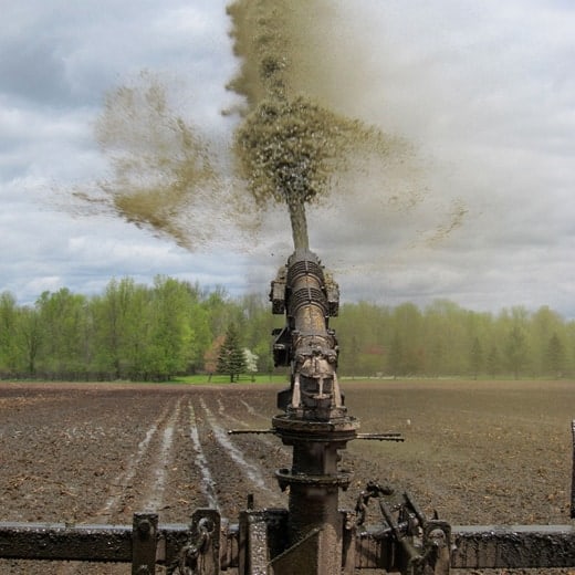 A large metal irrigation gun shooting a stream of liquid manure into the air over a field, creating aerosolized fecal particulate matter.