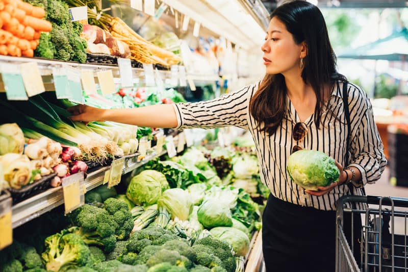 A woman grocery shopping in the produce section, along the perimeter of the store. She is holding a cabbage and reaching for a leek.