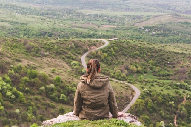 A woman sitting on a cliff's edge overlooking a curving road, symbolic of starting a new journey.