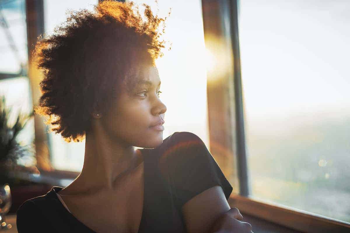 A young Black woman gazing out a set of windows with an aware but guarded look on her face as the sun illuminates her hair.