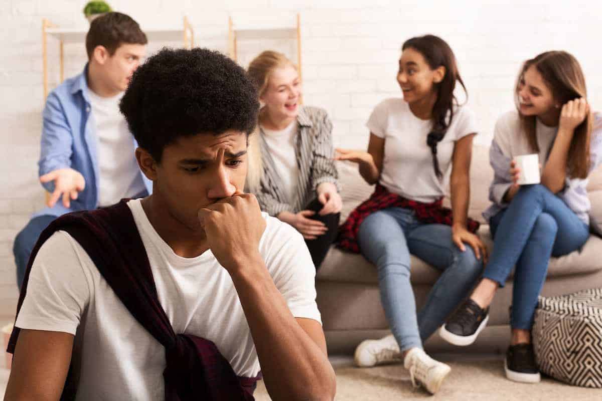 A Black teenager sitting apart from other teenagers socializing in the background, his forehead furrowed in distress from facing social challenges.
