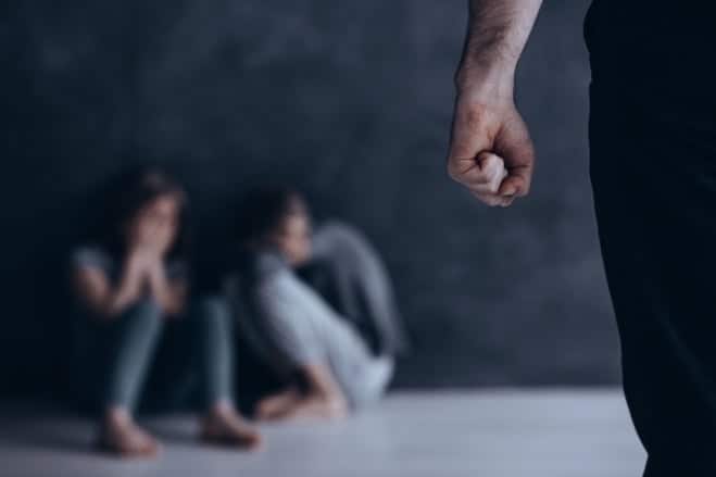 A man's clenched fist by his side as he stands in front of two figured huddled on the ground, representing a domestic violence situation.