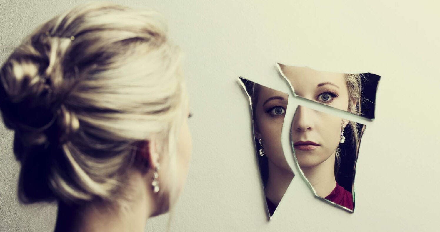 A White woman looks into a fractured mirror on the wall, reflecting her startled face broken into three pieces—representative of how vegans threaten our moral self-image.