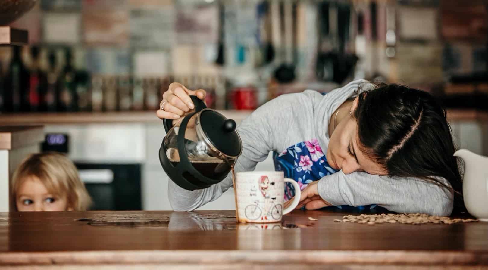 An exhausted woman lays her head down as she pours coffee onto the table, missing her mug entirely.