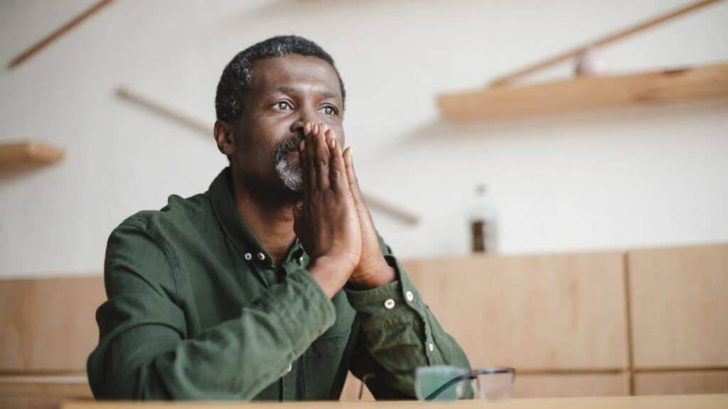 A Black man sits thoughtfully with his hands pressed together and held up to his mouth, in deep consideration.